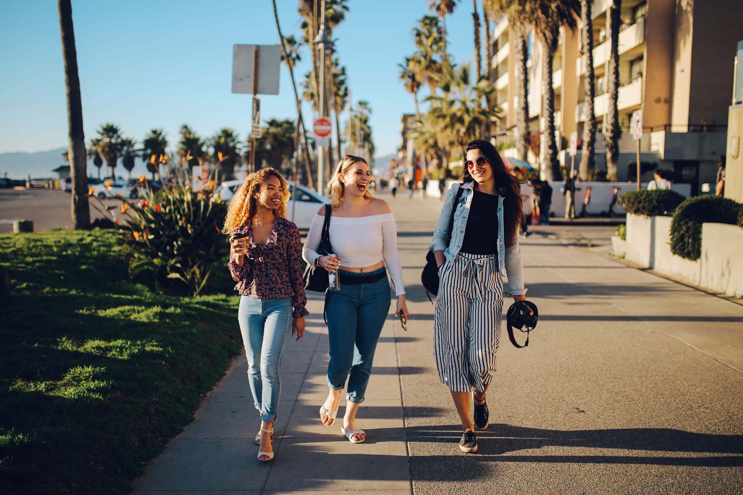 Three women walking down a sunny water front street, smiling and laughing.