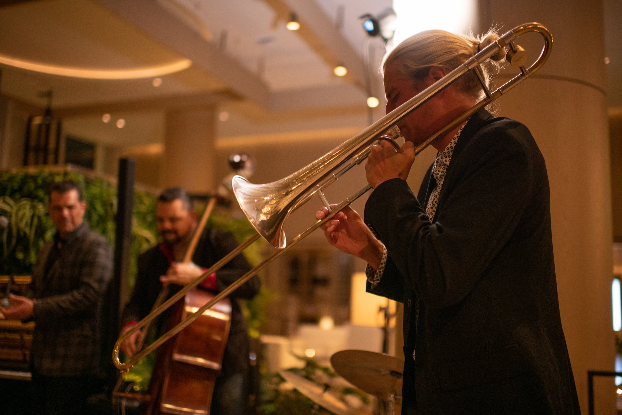 A man playing the trombone in a bar, a drummer and a cellist in the background.