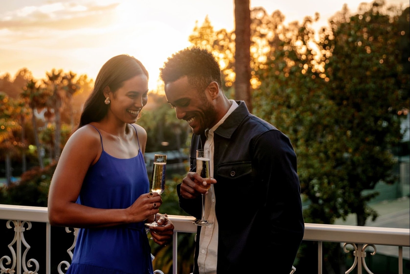 A couple laughing and holding flutes of champagne on a terrace at sunset.