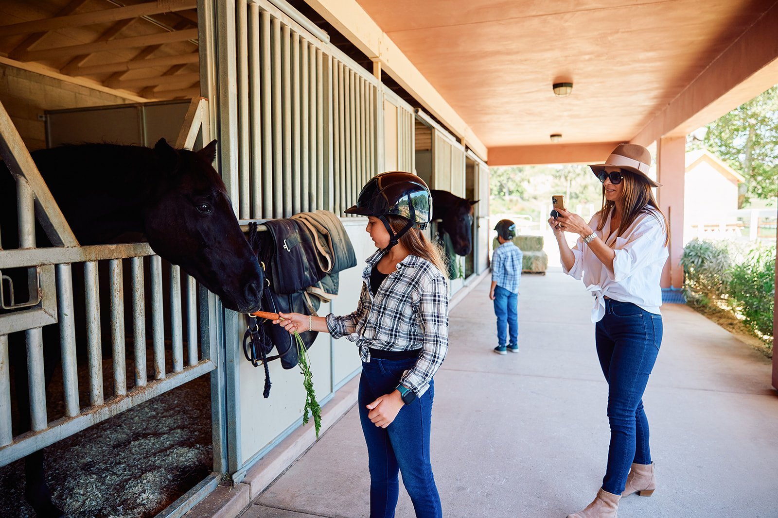 A young girl feeds a carrot to a horse in a stable while her mother takes a photo.