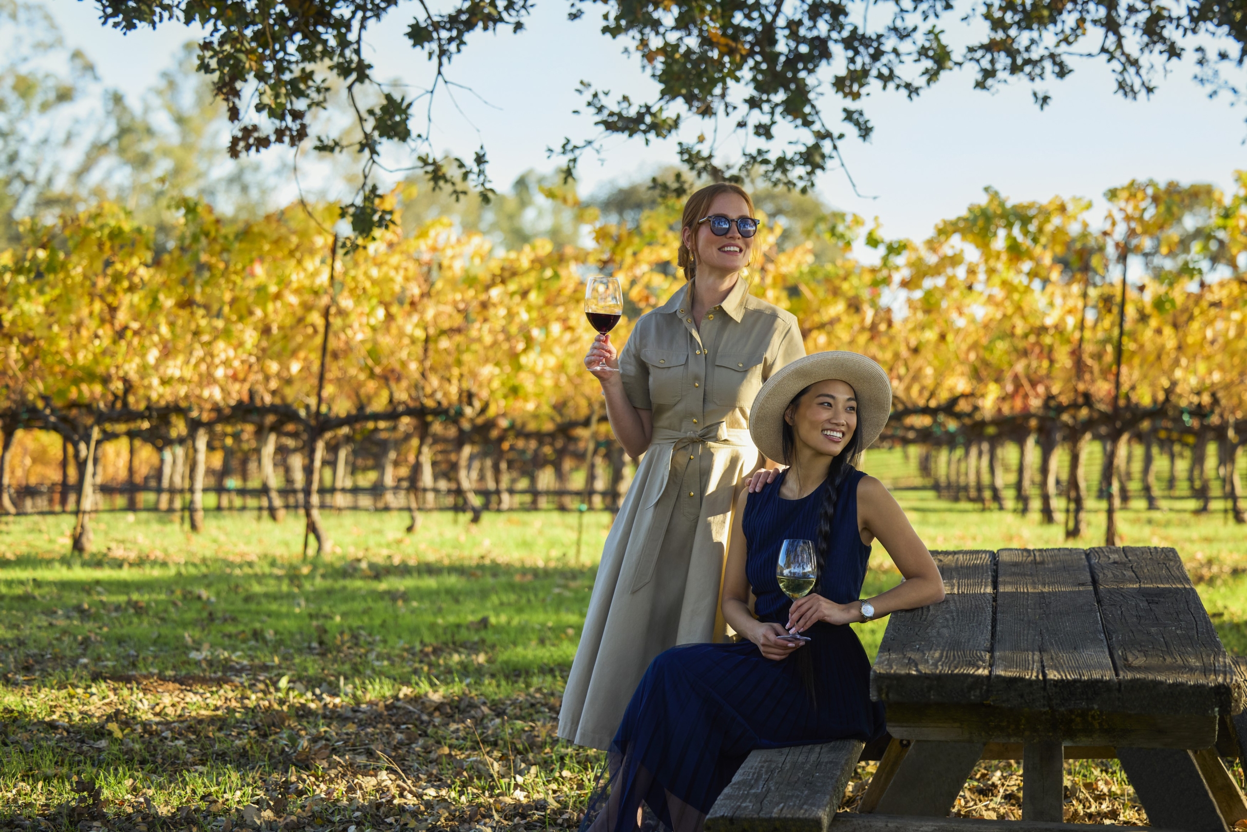 Two women in a vineyard sitting at a bench drinking wine.