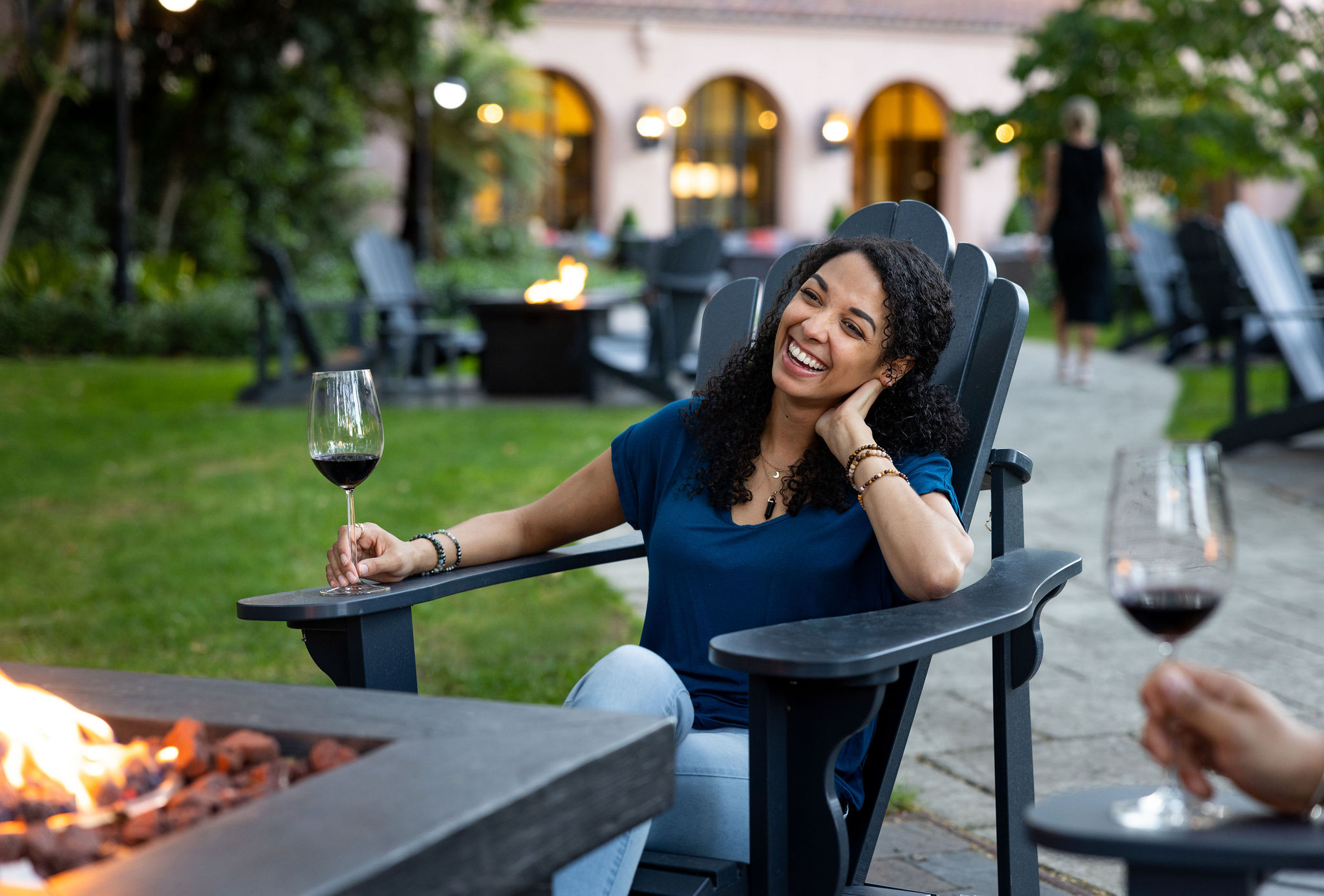 A woman smiling holding a glass of wine sitting in front of an outdoor fireplace.