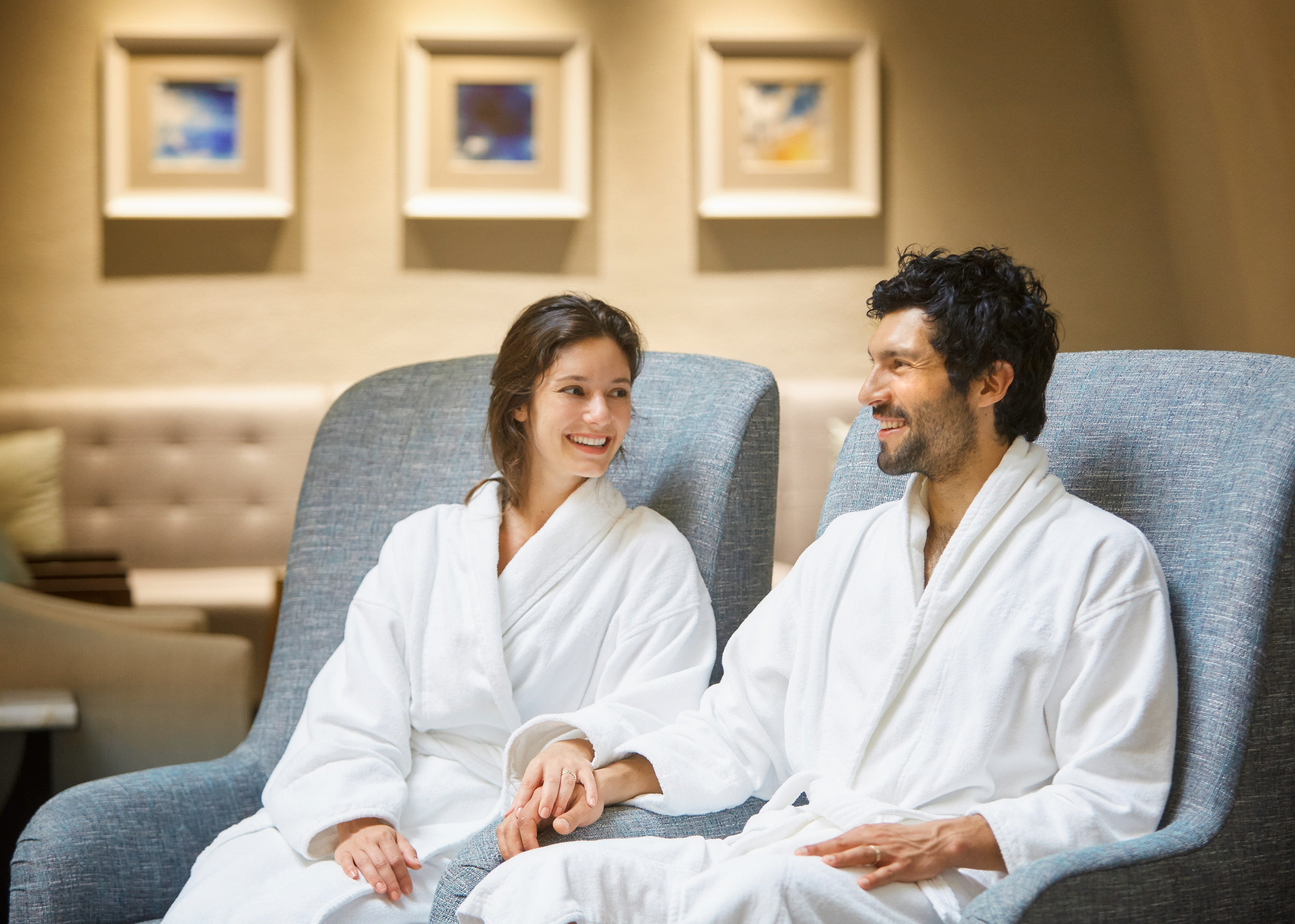 A couple sitting in spa chairs wearing spa robes and holding hands.