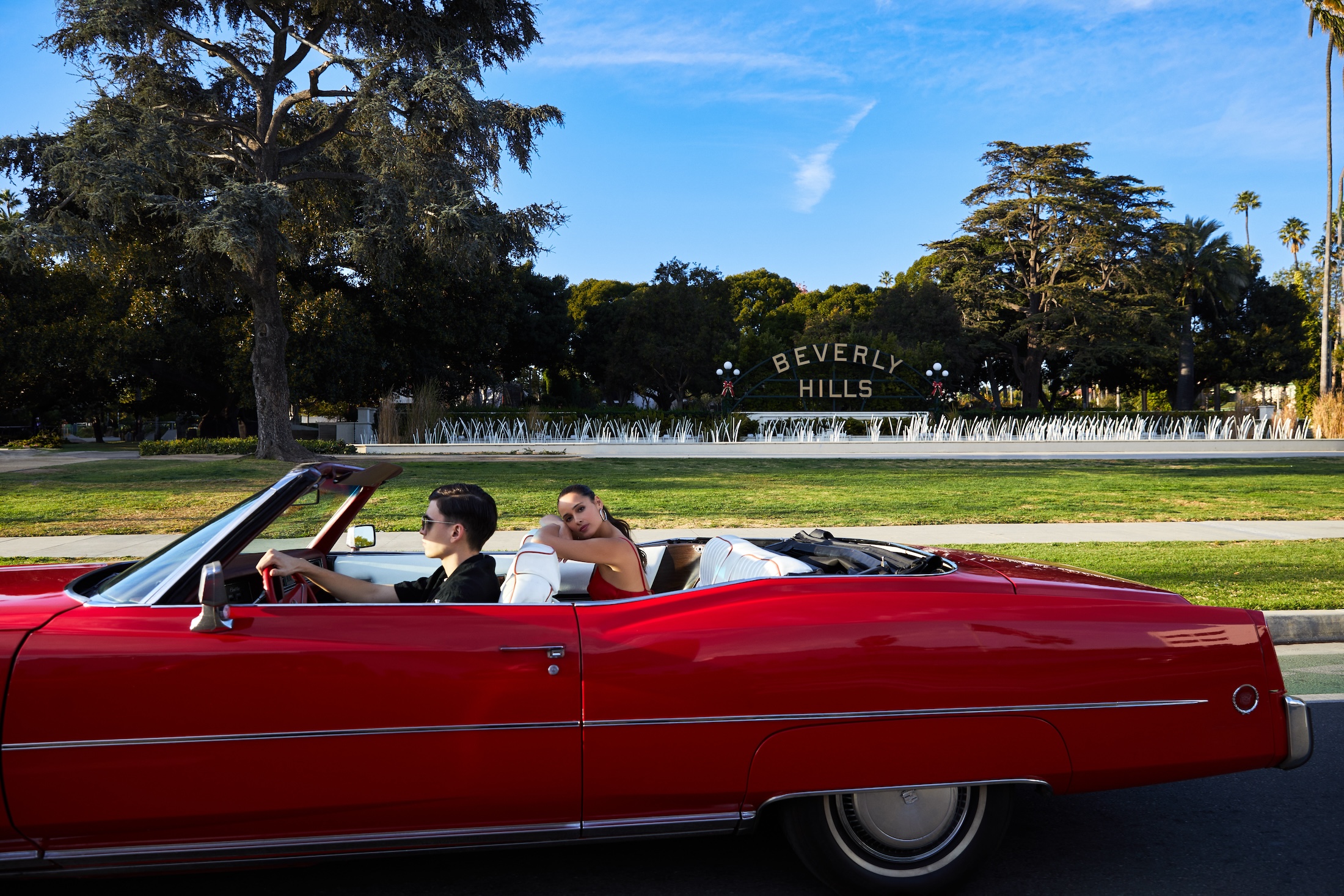 A young man driving a red convertible through Beverly Hills with a young woman in the back seat.